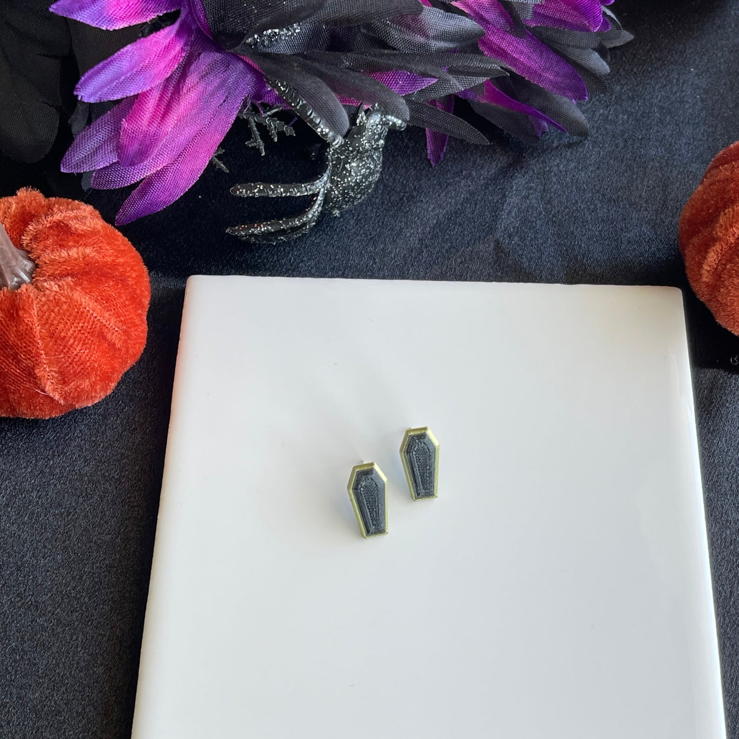 Pair of earrings on a white square platform with decorative pumpkins and flowers on a dark surface